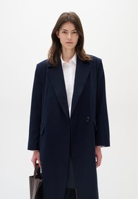 Cappotto lungo blu navy con chiusura a un pulsante, rever a sigaretta e tasche laterali, indossato sopra una camicia bianca. Borsa marrone in mano.