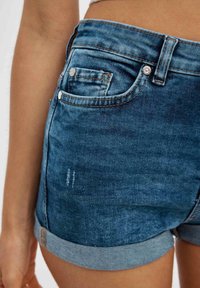 Pantalones cortos de mezclilla en azul oscuro con dobladillo deshilachado, puños enrollados y dos bolsillos traseros. Presenta un sutil desgaste y detalles de costura en contraste.