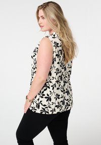 Mouwloze top in textuur crème stof met een zwart bloemenpatroon, voorzien van een ronde halslijn en een relaxed pasvorm. Gecombineerd met zwarte broeken.