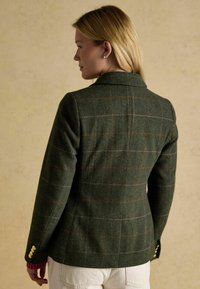 Vihreä villatakki, jossa on ruskea ja vaaleanharmaa ruutukuosi, yksittäinen takatuuletus ja kultaiset napit hihoissa. Teksturoitu kangas.