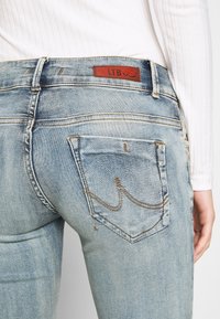 Jeans en denim clair avec une légère décoloration, ornés d'une étiquette en cuir rouge et de coutures décoratives sur les poches arrière.