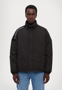 Sort polstret jakke med høj krave, lange ærmer og elastiske manchetter. Har logo på skulderen. Båret over blå jeans.