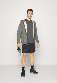 Grå zip-up hoodie med en cremefarvet inderside, mørk t-shirt, sorte shorts og sorte sneakers. Holder en klar vandflaske med sort låg.
