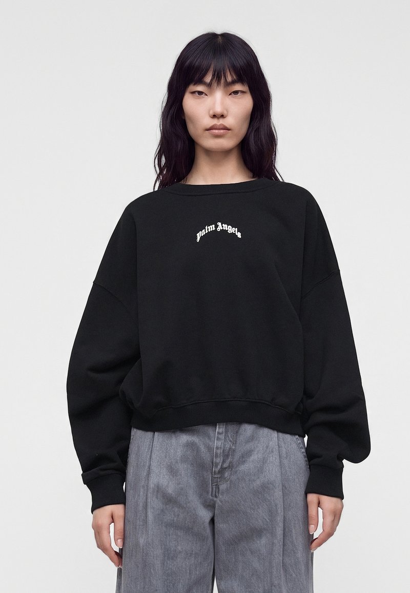 Νέος με μακριά μαύρα μαλλιά φορούν μαύρο φούτερ με λευκό κείμενο "palm angels" και γκρι φαρδιά παντελόνια, στέκεται μπροστά σε απλό φόντο.