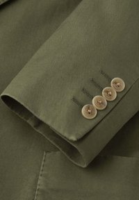 Ärmel eines olivgrünen, strukturierten Blazers mit vier beigen Knöpfen und sichtbaren Knopflöchern am Ärmelende, darunter ist ein Teil der Tasche zu sehen.