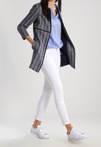 Giacca lunga a fantasia navy con colletto e tasche, abbinata a una camicia azzurra a bottoni, jeans skinny bianchi e sneakers bianche.