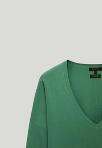 Pull en coton vert avec un col en V, une coupe décontractée et des manches courtes. Présente des bordures côtelées et une texture lisse. Étiquette visible à l'intérieur du col.