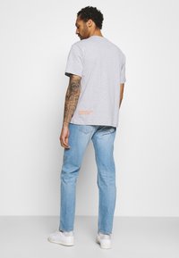 Camiseta de algodón gris con un corte relajado, cuenta con mangas cortas y un pequeño logo naranja en la parte trasera, combinada con jeans azul claro y zapatillas blancas.