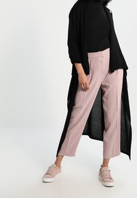 Zwarte lange cardigan over een zwarte coltrui, gecombineerd met lichtroze geplooide broek en lichtroze sneakers. Eenvoudig ontwerp met zachte texturen.