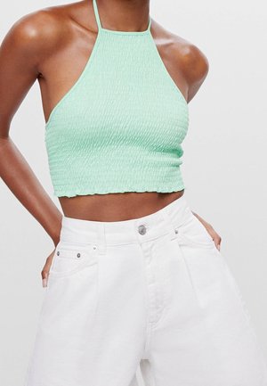 Woman wearing a mint green textured halter crop top and high-waisted white pants with hands resting in pockets.