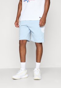 Light blue athletic shorts with white side panel and logo, paired with white sneakers and socks. The model wears a plain white t-shirt.