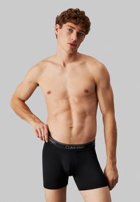 Crne bokserice s Calvin Klein logom na pojasu, koje nosi model bez majice s kovrčavom kosom, na sivoj pozadini.