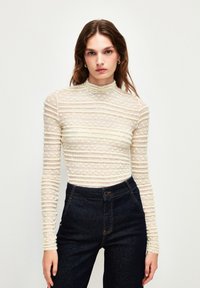 Hvid, langærmet bluse med høj hals, der har et struktureret stribet mønster. Sammen med mørkeblå højtaljede jeans.
