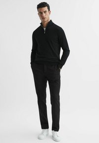 Pulover negru cu fermoar, confecționat dintr-un material neted, asociat cu o pereche de pantaloni negri tapered. Adidași albi completează ținuta, creând un look monocrom.