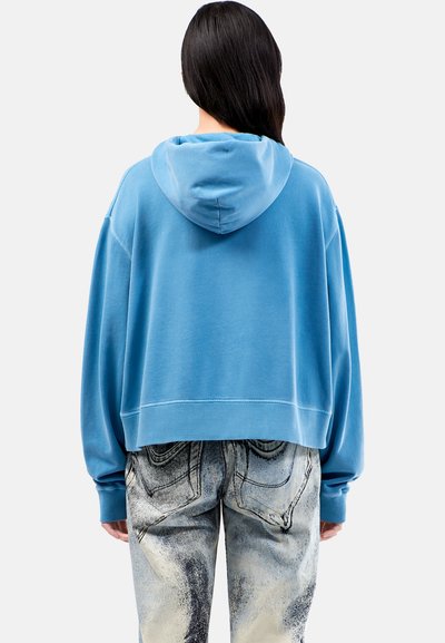 Felpa azzurra tagliata, con cappuccio regolabile e polsini a coste, abbinata a jeans di denim grigio chiaro a fantasia con dettagli sulle tasche.