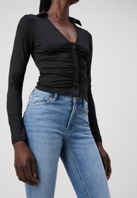 Person som bär en figurnära svart långärmad skjorta med knappar och högmidjade blå jeans mot en enkel ljus bakgrund.