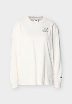 Chemise blanche à manches longues et col rond avec un petit texte noir "ADIDAS three stripes STUDIO" sur la poitrine gauche et le logo Adidas sur le poignet droit.