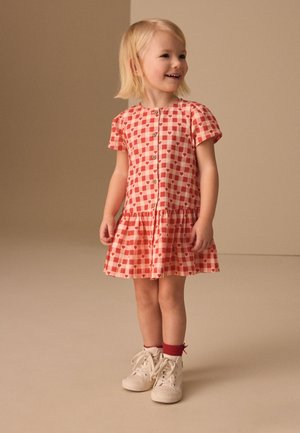 Fiatal gyermek mosolyog, áll, rózsaszín és piros szívmintás ruhát, fehér sportcipőt és piros zoknit visel egyszínű bézs háttér előtt.