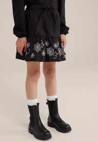 Zwarte tiered rok met witte bloemenborduring aan de zoom, gecombineerd met zwarte enkellaarsjes en witte sokken. Heeft een gestrikt tailleband.