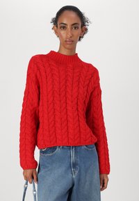 Rød strikket genser med flettemønster, rund hals og lange ermer. Kombinert med lys blå denim.