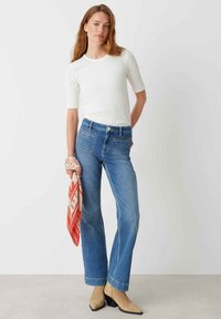 Witte geribbelde top met korte mouwen, gecombineerd met hoge blauwe jeans. Model houdt een rode en witte sjaal met patroon vast en draagt beige hakken.
