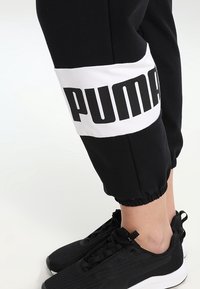Zwarte joggingbroek met een witte band waarop "PUMA" in vetgedrukte zwarte letters staat gedrukt. De boorden zijn elastisch, gecombineerd met zwarte sportschoenen.