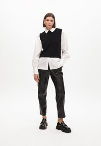 Schwarzer ärmelloser Pullover über einem weißen Hemd mit Kragen, kombiniert mit schwarzen Lederimitat-Hosen und klobigen schwarzen Schuhen. Glatte Texturen und taillierter Schnitt.