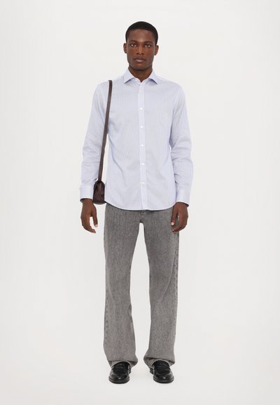 Camicia a maniche lunghe di colore azzurro chiaro con strisce bianche, abbinata a pantaloni grigi a gamba larga e scarpe nere, mentre tiene una piccola borsa a tracolla marrone.