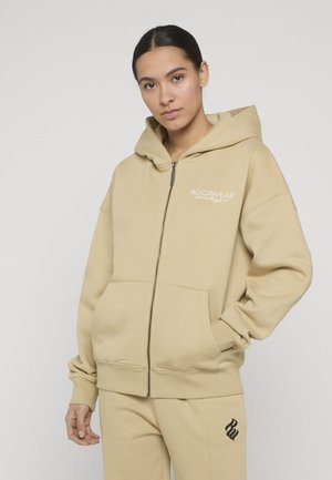 Jeune femme portant un sweat à capuche zippé beige Rocawear et un pantalon assorti, les mains dans les poches, debout devant un fond uni.