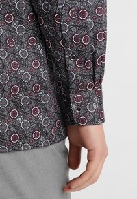 Dlouhý rukáv košile s tmavým vzorovaným materiálem s kruhovými motivy v červené a bílé barvě, rukáv s knoflíkovým zapínáním a kontrastní šedé kalhoty.