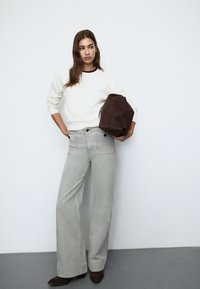 Svetlo sivi široki traperici z velikimi žepi, v kompletu s smetano svetrom. Temno rjav sueden torbica je držana pod pazduho.