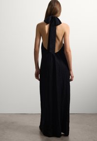 Abito lungo nero con scollo a halter, realizzato in tessuto testurizzato, schiena scoperta e grande fiocco al collo, caratterizzato da una silhouette fluida.