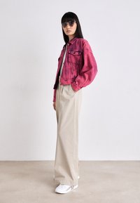 Giacca di jeans rosa con tasche frontali, abbinata a pantaloni larghi beige e sneakers bianche. Il modello indossa occhiali da sole rotondi scuri.