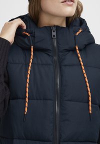 Marinblå puffer väst med hög krage, prydd med en orange dragsko och en framzipper. Kviltad textur med kontrasterande detaljer.