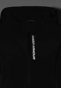 Čierna bunda s kapucňou a predným zipsom, na pravej strane s logom "UNDER ARMOUR" v bielej farbe. Hladká textúra a priliehavý strih.