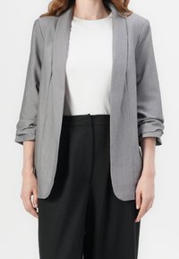 Blazer gris à la texture douce, au design ouvert et aux manches retroussées, associé à un t-shirt blanc et un pantalon noir pour un contraste marqué.
