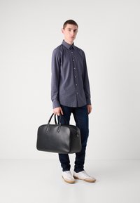 Borsa da viaggio nera in pelle con angoli arrotondati, manici doppi e chiusura con zip. Il modello indossa una camicia blu a fantasia e jeans scuri con sneakers bianche.