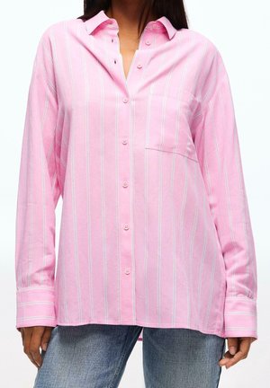 Femme portant une chemise rose à boutons avec des rayures verticales blanches et un jean bleu, montrée du cou aux hanches sur fond blanc.