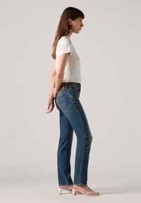 Donkerblauwe jeans in een recht model met een klassieke vijf-pocketontwerp. Gecombineerd met een lichtgrijze t-shirt en witte sandalen met bandjes.
