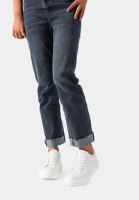 Sneakers bianchi in tessuto testurizzato, con punta tondeggiante e sei occhielli, abbinati a jeans in denim blu arrotolati ai polsini per un look casual.