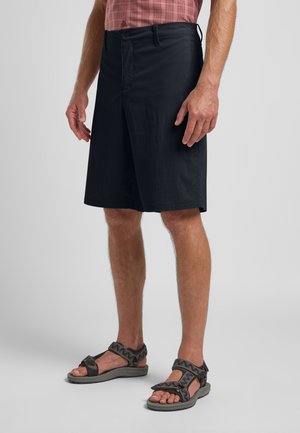Schwarze Shorts aus leichtem Stoff, mit flachem Schnitt, Seitenentaschen und klassischer Passform, kombiniert mit dunklen Sandalen mit verstellbaren Riemen.