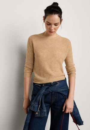 Femme portant un pull beige à manches longues avec une veste en jean nouée autour de la taille, regardant vers le bas sur un fond uni.