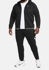 Sudadera negra de Nike con cremallera y bolsillos frontales, combinada con joggers negros que tienen un logo blanco y zapatillas deportivas blancas.