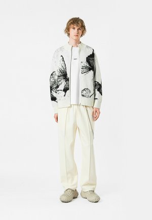 Chaqueta bomber de punto con gráfico de mariposa en blanco y negro, combinada con una camiseta blanca y pantalones anchos color crema, completada con zapatos beige texturizados.
