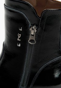 Botte chevillère en cuir noir avec fermeture éclair sur le côté, texture brillante, clous en argent décoratifs et surpiqûres contrastantes le long des coutures.