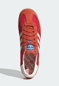 Punainen Adidas-tenniskenkä, jossa on valkoiset raidat, samettinen kärki ja sininen logo-lappu kielekkeellä, nähtynä ylhäältä vaalealla taustalla.