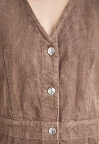 Abito in corduroy marrone con scollatura a V profonda, caratterizzato da cinque bottoni a pressione argentati sul davanti e una cintura di stoffa in vita. Tessuto testurizzato.