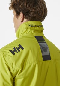 Chaqueta amarilla impermeable con acentos de logo en negro, cuello alto y panel trasero reflectante para mayor visibilidad. Tejido suave y ligero.