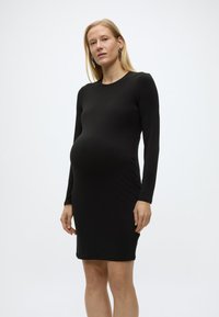 Έγκυος γυναίκα με μακριά ξανθά μαλλιά, ντυμένη με εφαρμοστό μακρυμάνικο μαύρο φόρεμα, που στέκεται μπροστά σε απλό λευκό φόντο.