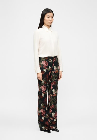 Nő áll, hosszú ujjú krémszínű blúzt visel, amelyet fekete, széles szárú nadrágba tűrt, nagy rózsaszín és piros virágmintákkal díszítve.